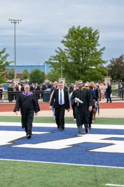 Graduates in caps and gowns walking on a football field during a graduation ceremony.