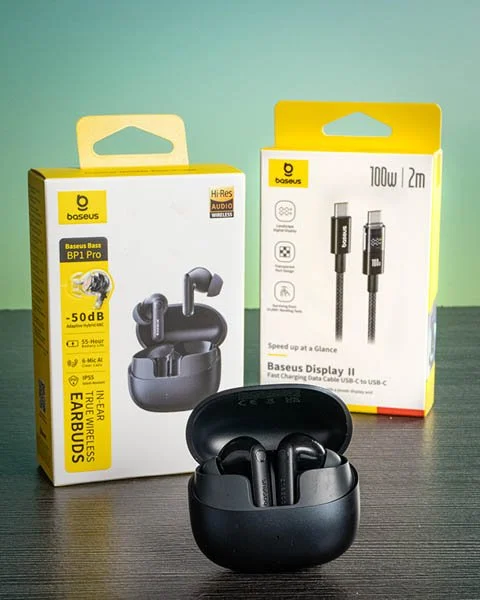 Wireless earbuds in a charging case with packaging boxes behind them, one for earbuds and the other for a USB-C charging cable, on a dark surface against a green background.