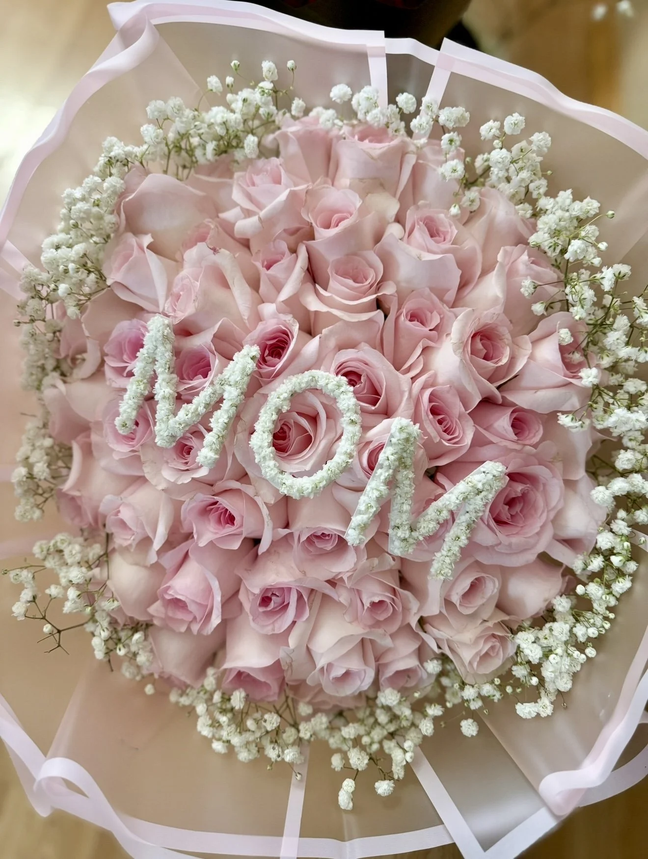 A bouquet of pink roses with white baby's breath flowers, featuring the word 'MOM' spelled out with white floral-covered letters.