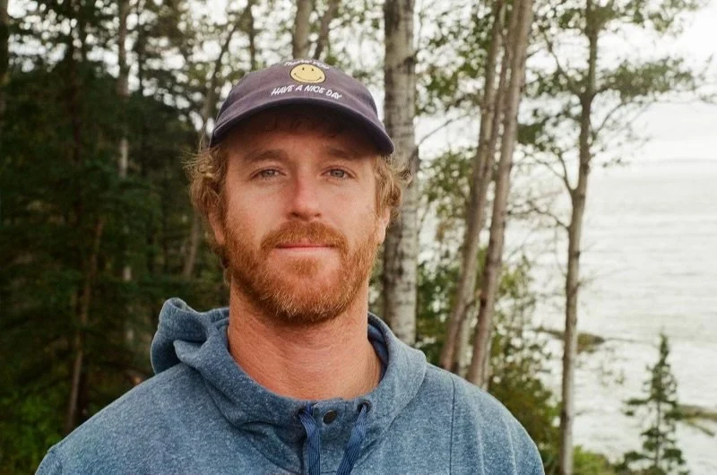 A man with a beard and curly hair wearing a blue hoodie and a black cap with a yellow smiley face and the words 'Have a Nice Day' printed on it, standing outdoors in front of trees and a body of water.