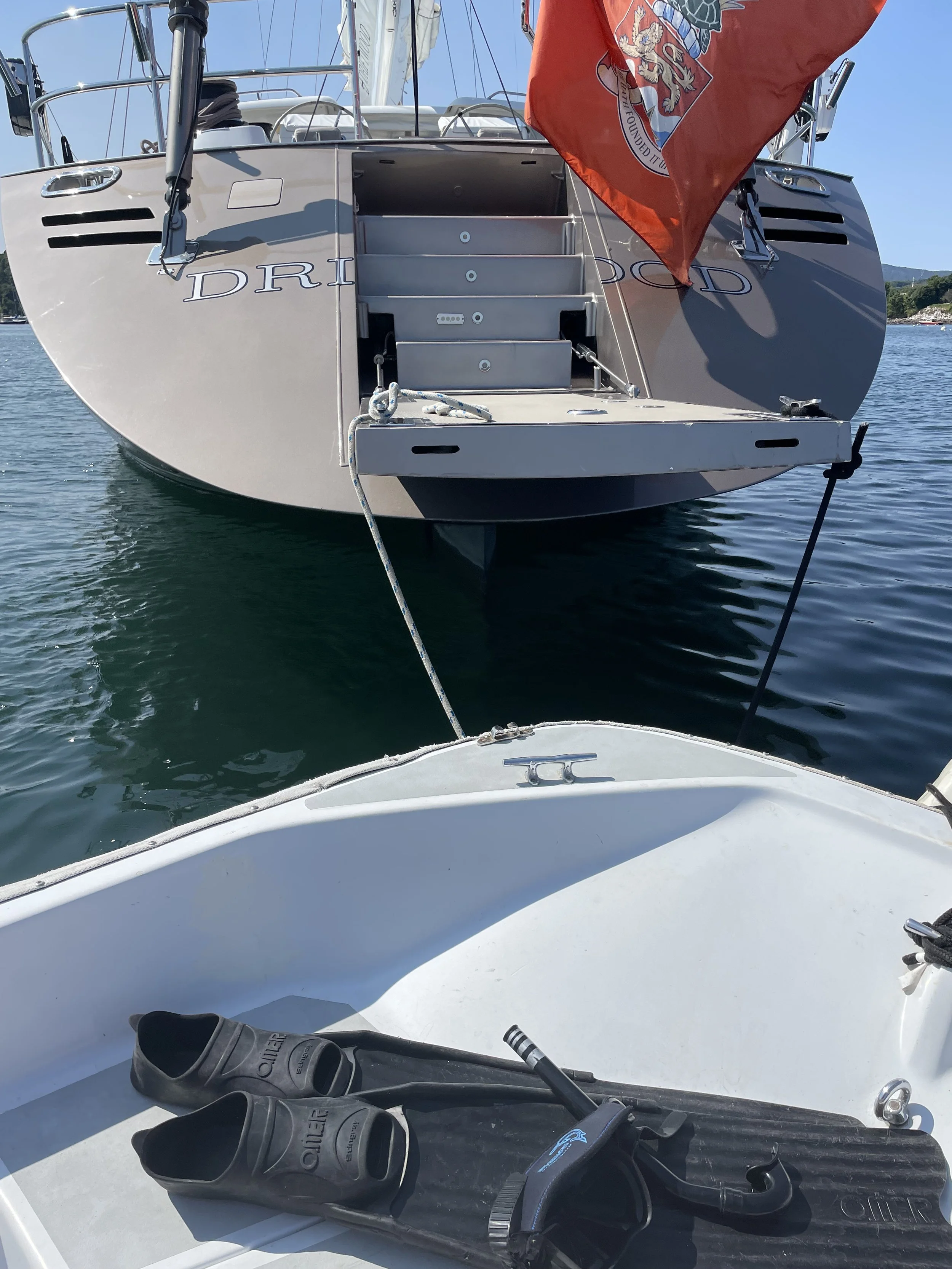 View of the rear of a white sailing yacht with steps leading up to the deck, a red flag with a crest flying on the right, and the calm water and shoreline in the background. The foreground shows a boat's dashboard with black rubber water shoes and a