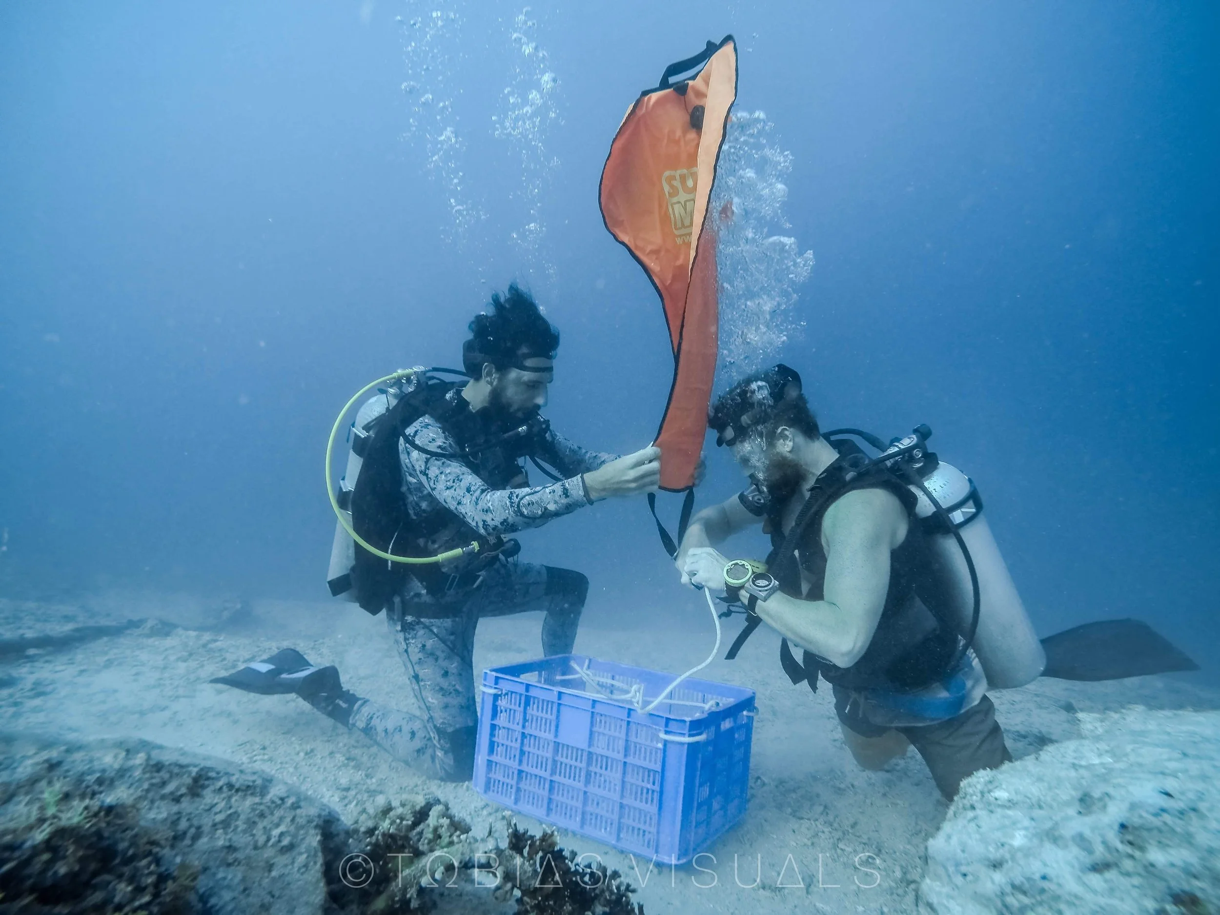 Two scuba divers underwater exchanging or inspecting an orange bag near a blue basket, with coral and rocks on the ocean floor.