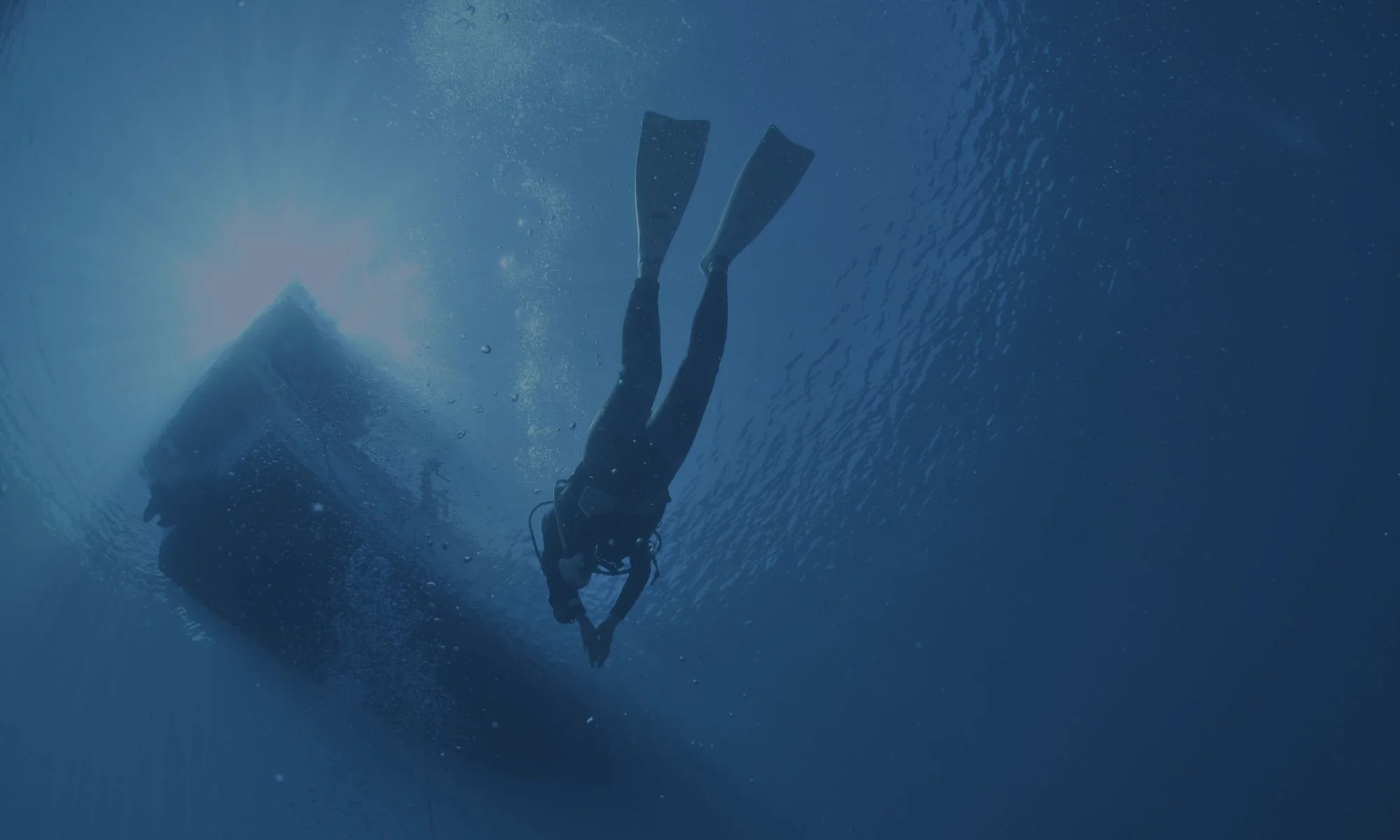 A person scuba diving underwater, holding a camera and surrounded by blue water with bubbles and sunlight filtering through.