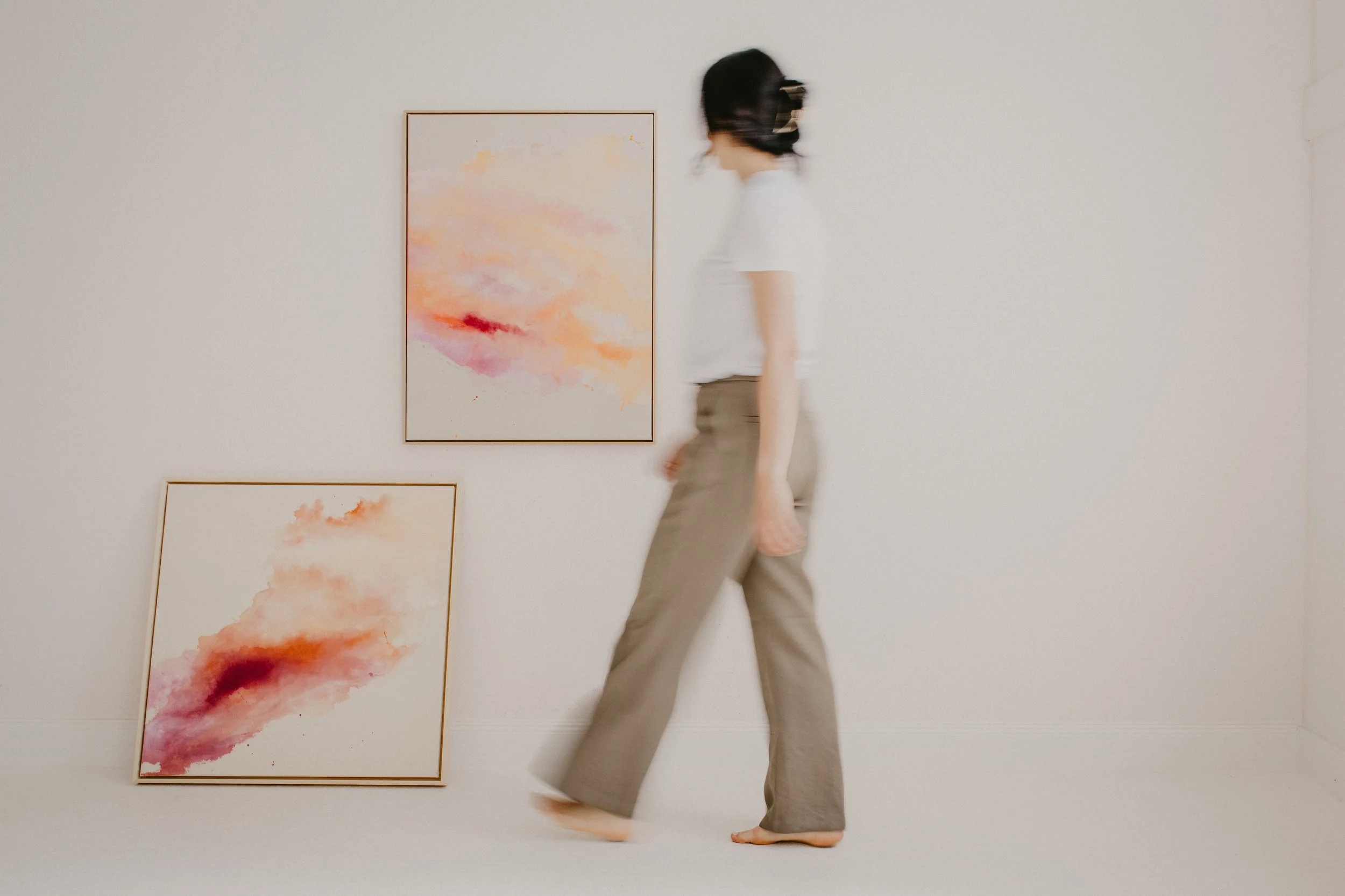 Eine Frau in hellen Hosen und ohne Schuhe, die von hinten geht, steht vor einer weißen Wandmit mehreren abstrakten Gemälden in Pastellfarben, die an der Wand hängen.