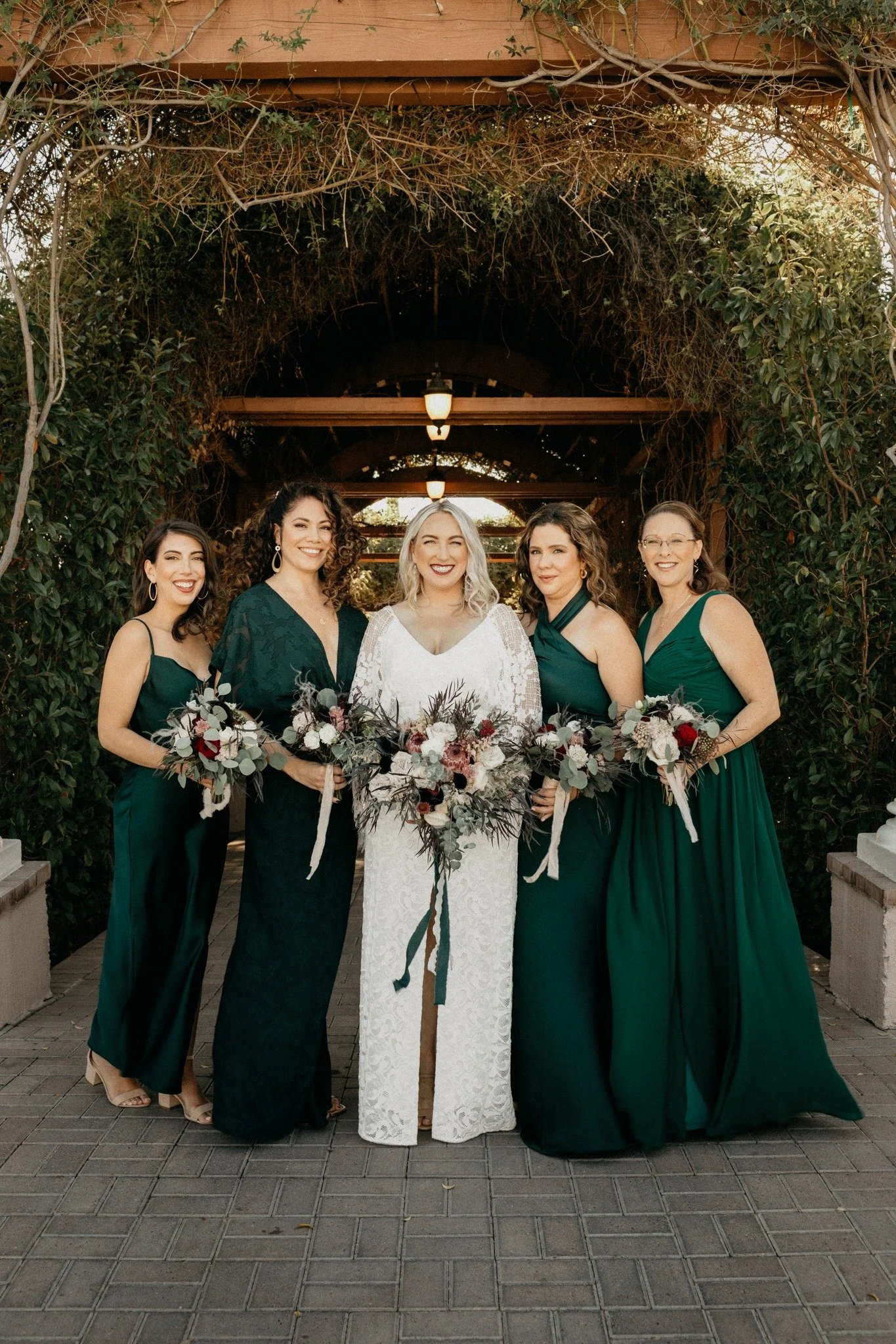 Bride in white gown posing with four bridesmaids in dark green dresses, each holding floral bouquets, standing under a vine-covered archway in an outdoor garden setting.