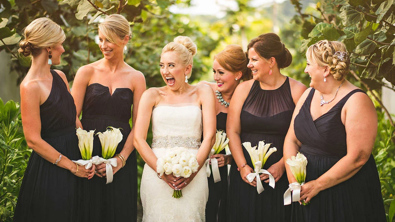 A wedding scene with five women, including a bride in a white dress holding a bouquet, and four bridesmaids in black dresses, all smiling and standing outdoors surrounded by green foliage.