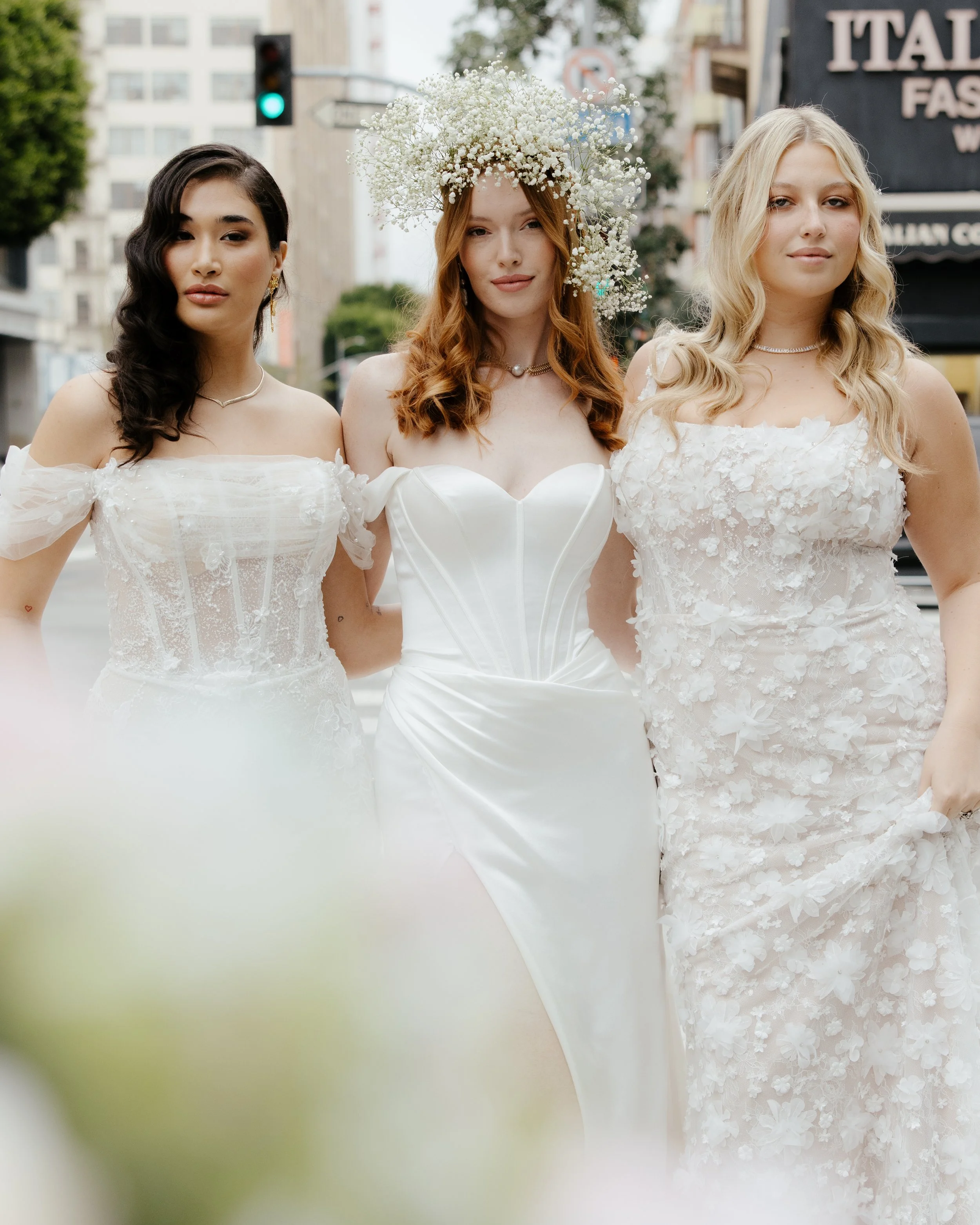 Three women in wedding dresses standing outdoors on a city street, with buildings and traffic lights in the background.