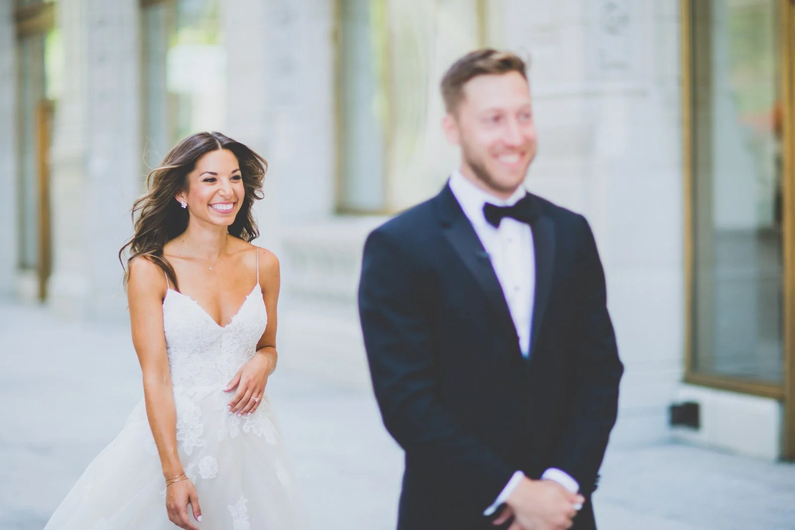Bride in a white wedding dress smiling and looking at groom in a black tuxedo, who is facing away from the camera, in an outdoor urban setting.