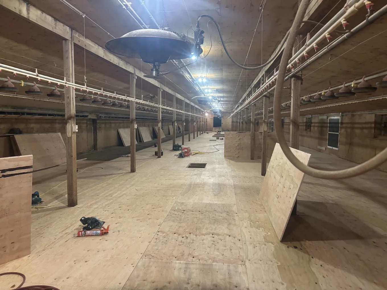 Long indoor farm or greenhouse under construction with wooden flooring, hanging irrigation or sprinkler system, and unfinished wooden walls. There are construction tools and materials scattered on the floor.