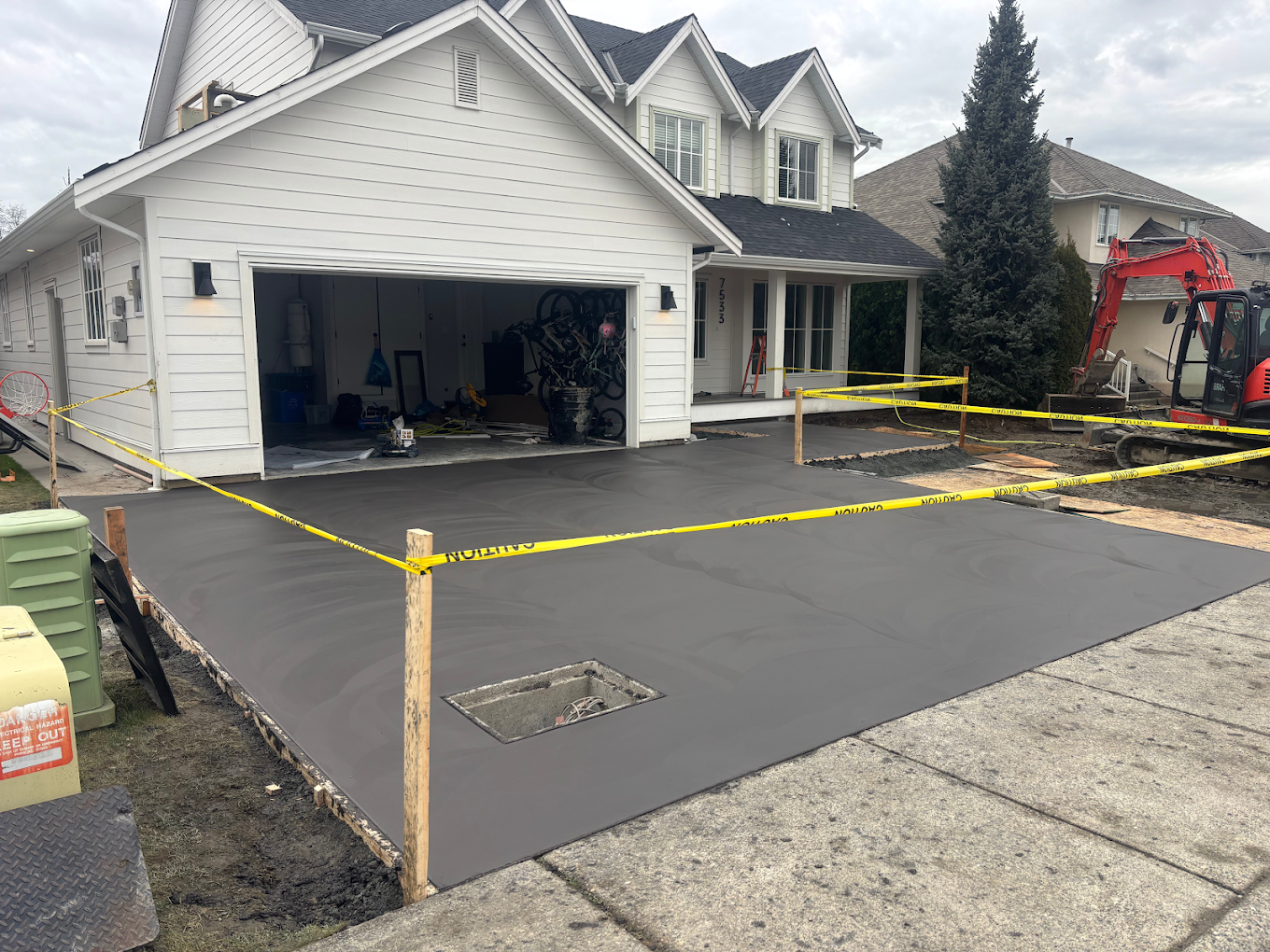 Newly poured concrete driveway in front of a white house with black shingled roof, yellow caution tape around the concrete, and construction equipment such as an orange mini excavator nearby.