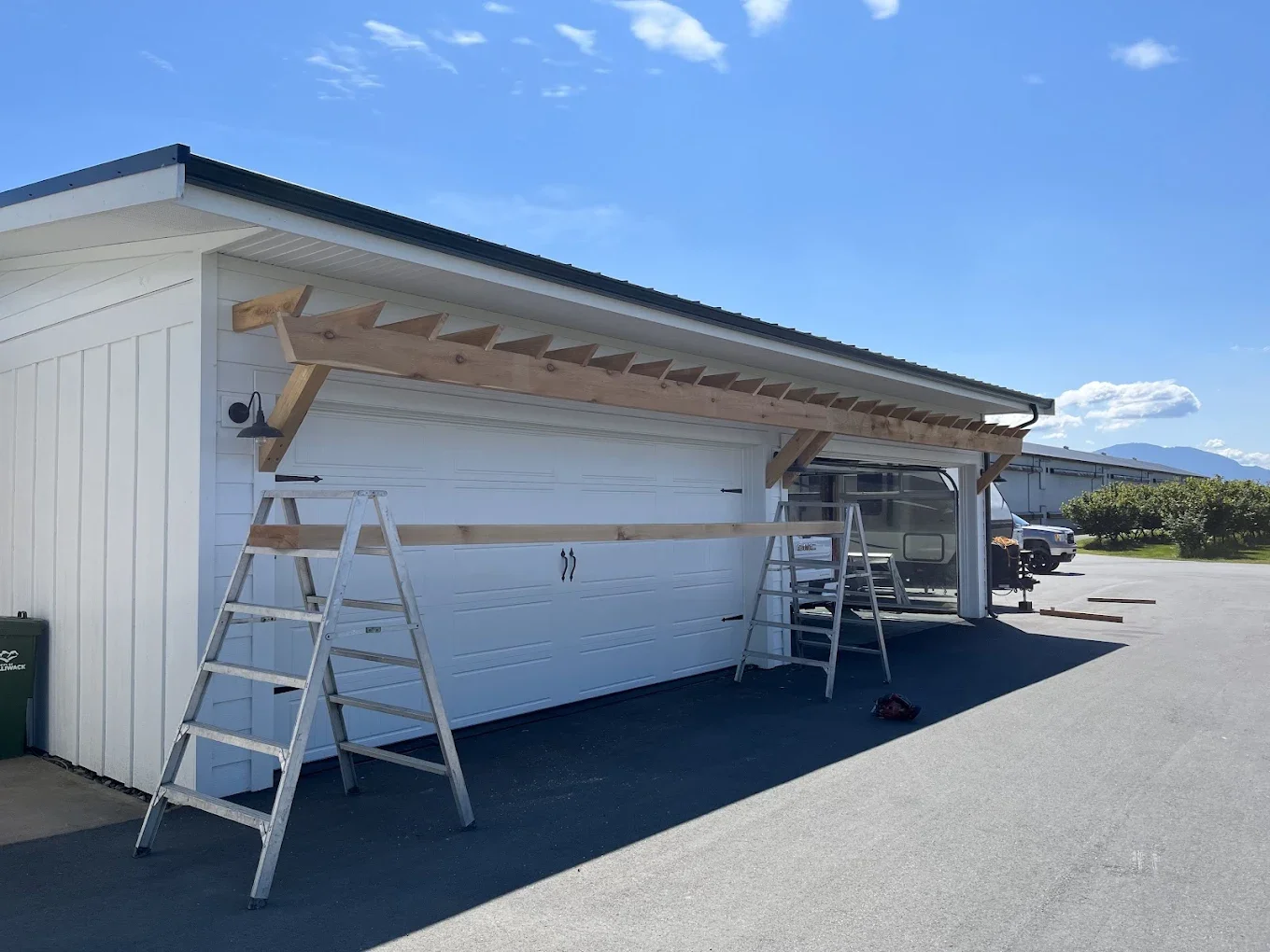 Garage door with wooden support beams being installed, ladders and construction tools nearby, residential area with parked vehicles and mountains in the background.