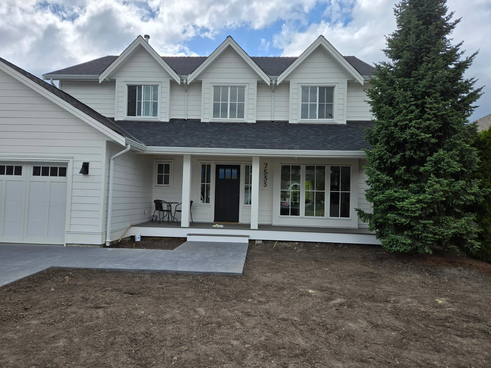 Newly constructed white two-story house with black door, large front window, garage on the left, and a large evergreen tree on the right, with bare dirt yard in front.
