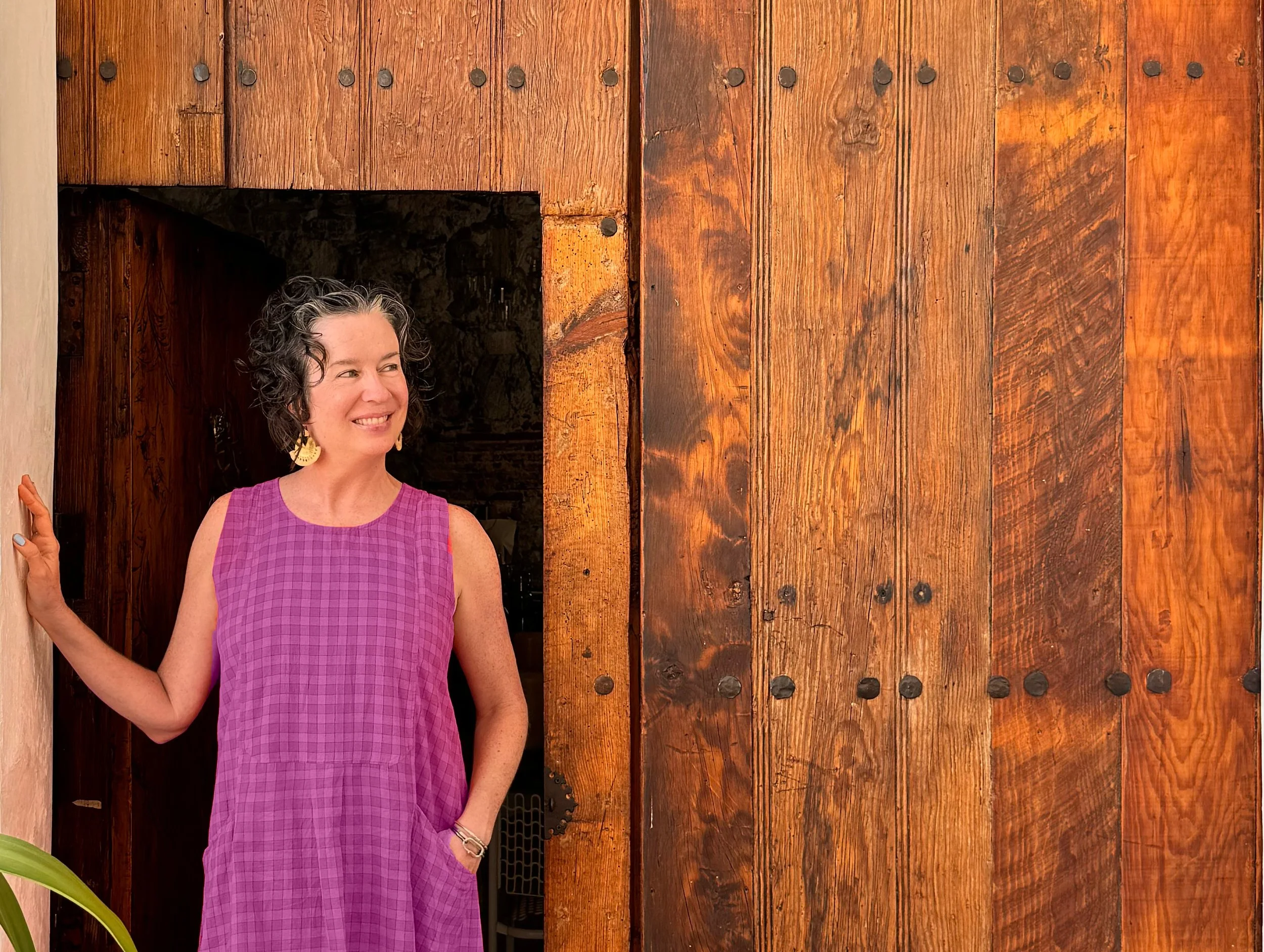 A woman with curly gray hair, wearing a sleeveless pink checkered dress, smiling and standing by a wooden door.