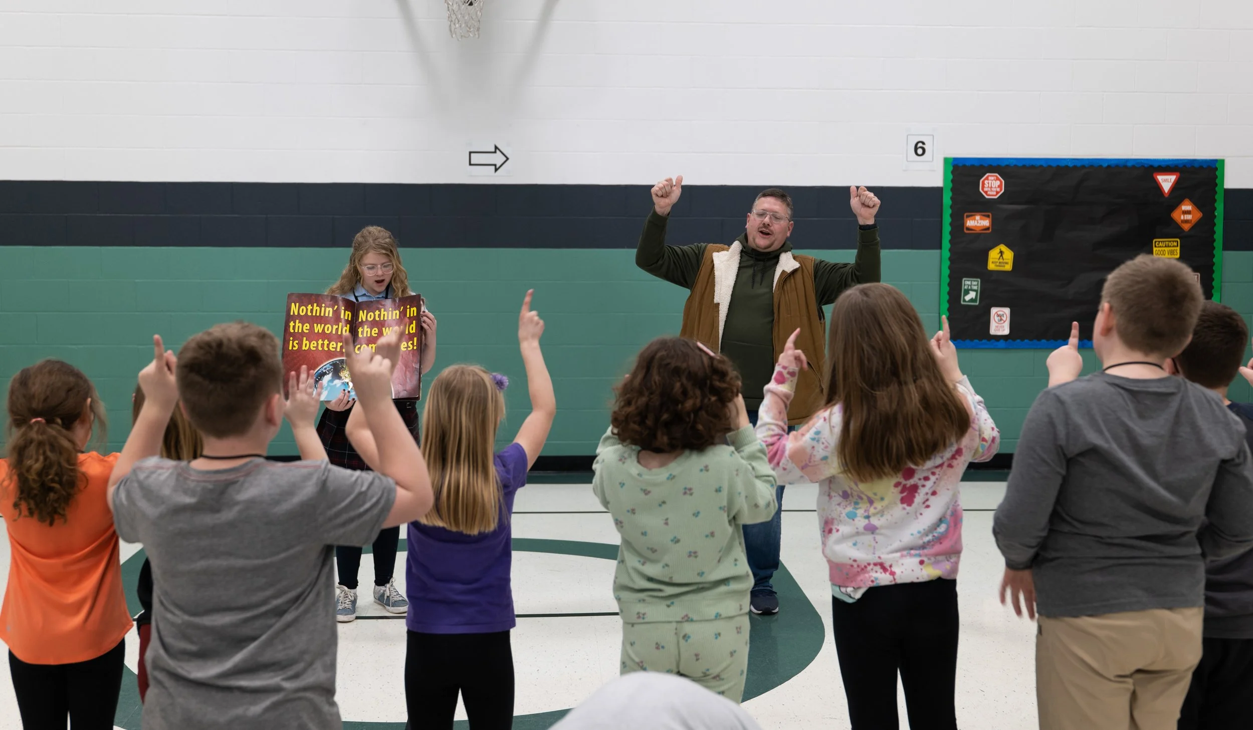 A group of children in a gymnasium playing a game of Simon Says, with a man leading the game, holding a sign that says "Nothin' in the world is better" and the children raising their hands.
