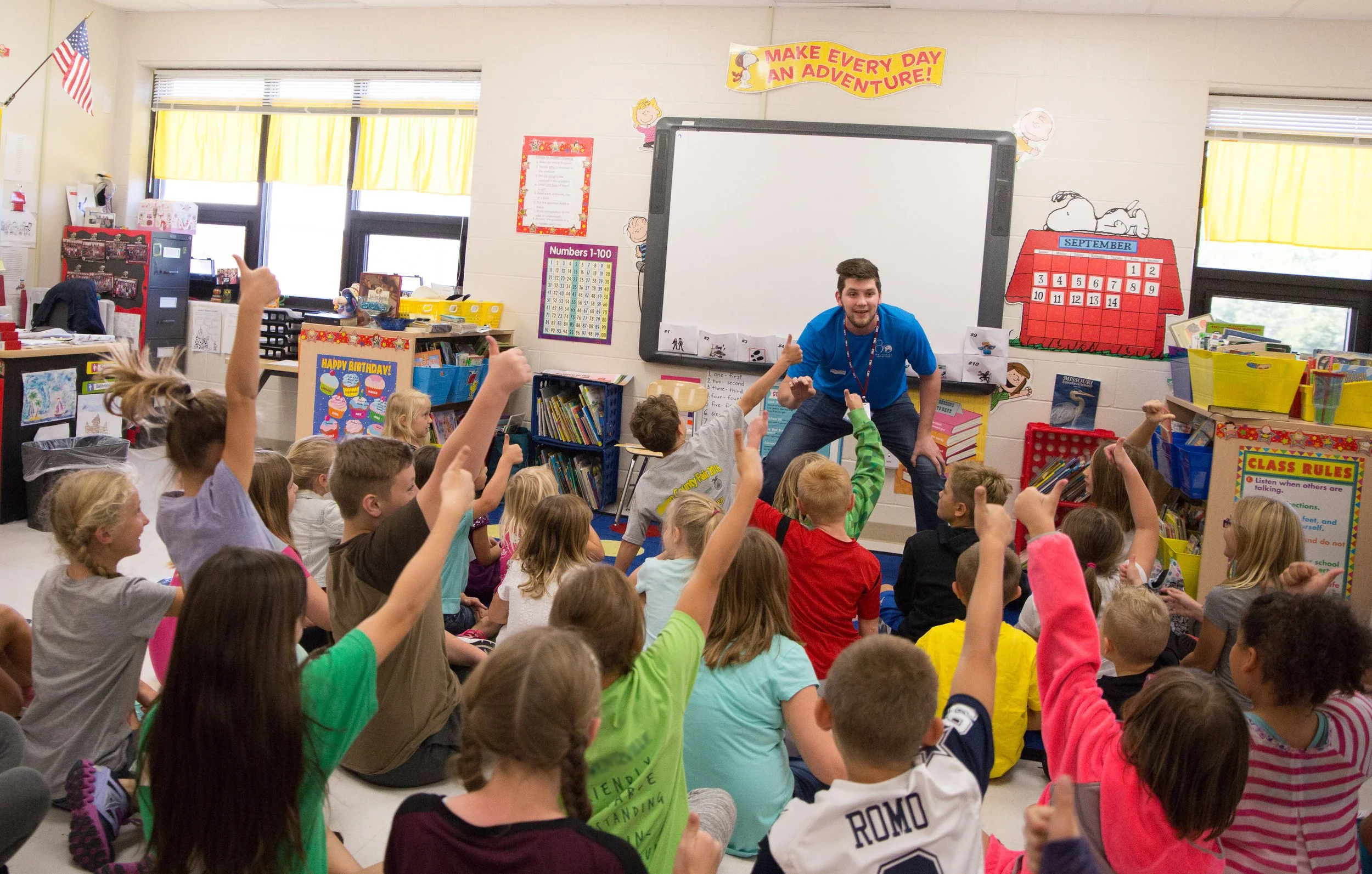 Elementary school classroom with young students sitting on the floor raising hands, a teacher at the front near a whiteboard, colorful decorations, and educational posters.