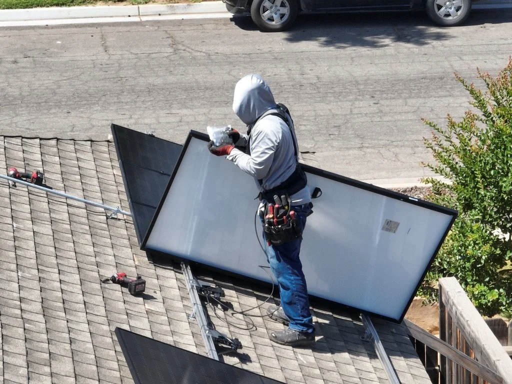 A worker installing or repairing outdoor solar panels on a roof during daytime, wearing a hooded jacket and gloves, with tools attached to his belt.