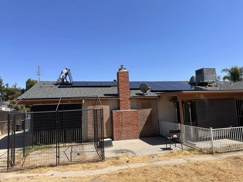 Back view of a house with solar panels on the roof, brick chimney, satellite dish, air conditioning unit, and a fenced yard with a chair and a white railing.
