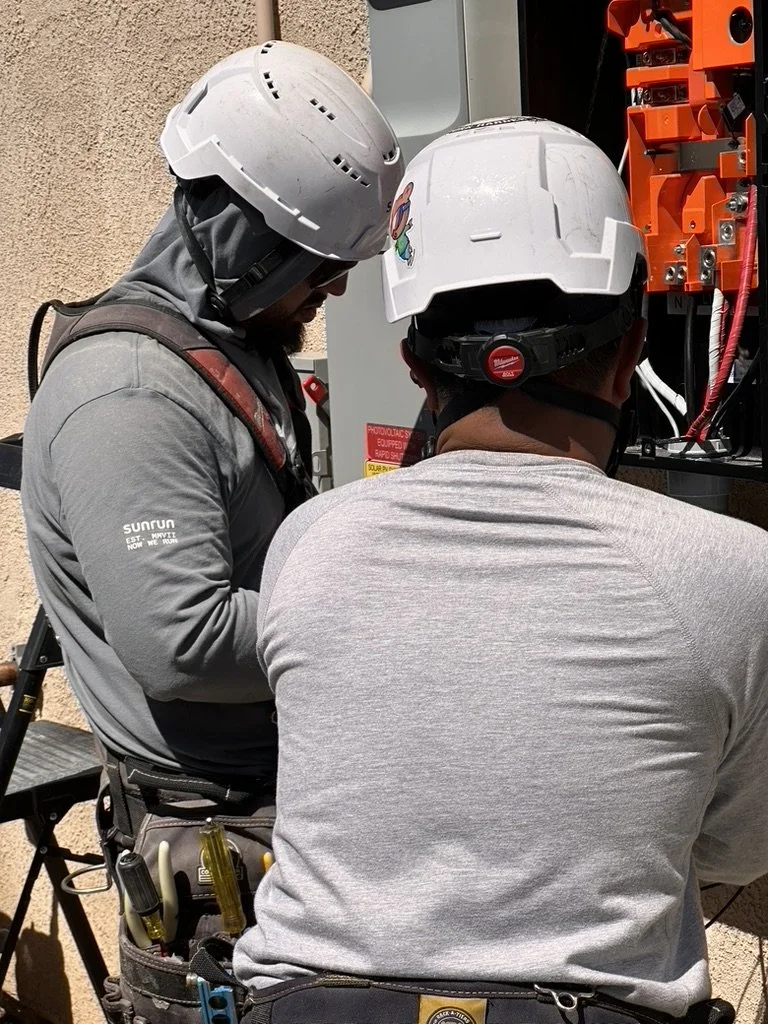 Two electricians wearing white hard hats working on an electrical panel outdoors, with tools attached to their tool belts.