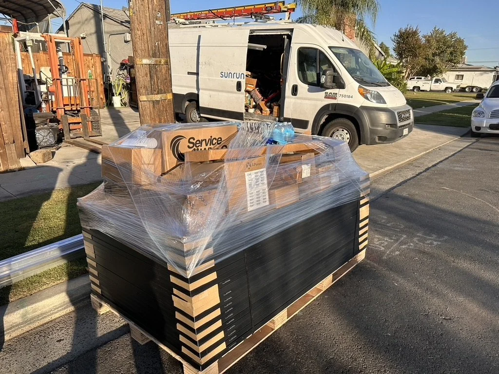 A pallet with boxes and equipment wrapped in plastic, placed in a parking lot near service vehicles and vehicles, with a wooden fence and trees in the background.