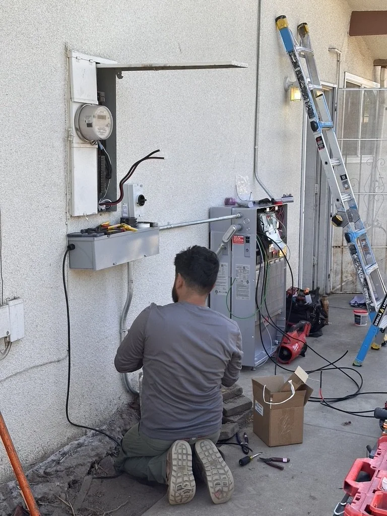 A man working on electrical or HVAC equipment outside, kneeling on the ground with tools and a cardboard box nearby, a ladder leaning against the wall, and various electrical components and wires.