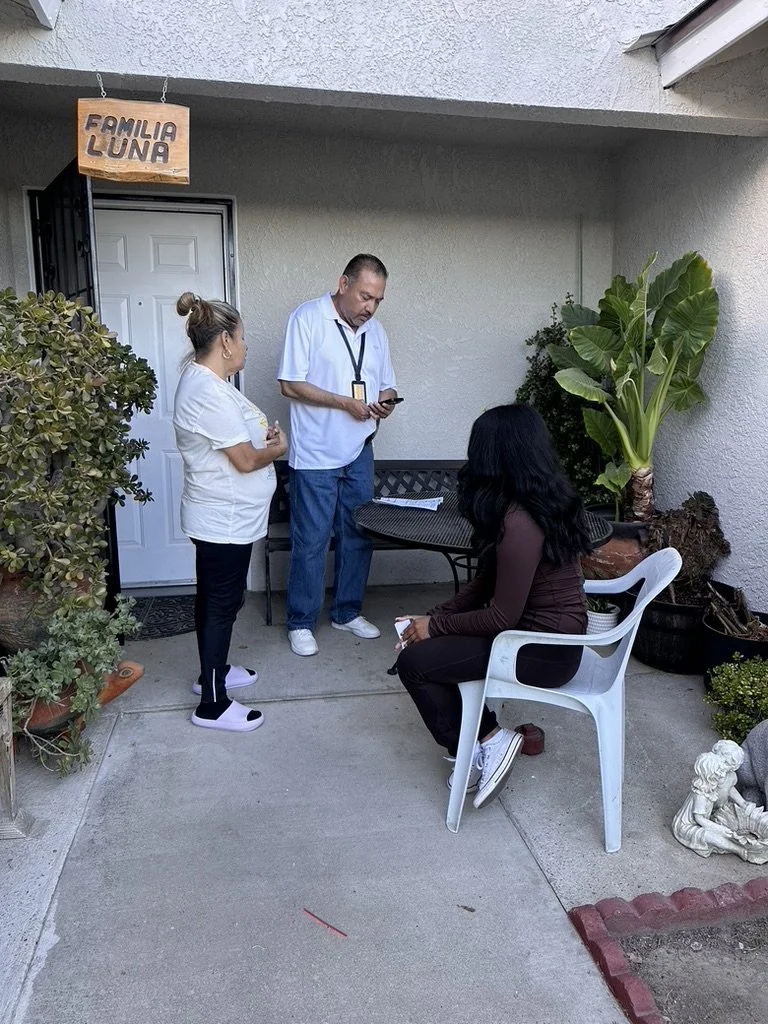 Three people are gathered outside a house with a small sign that reads "Familia Luna." One woman is standing, another woman is seated on a white outdoor chair, and a man is standing is looking at his phone. There are potted plants and decorative item
