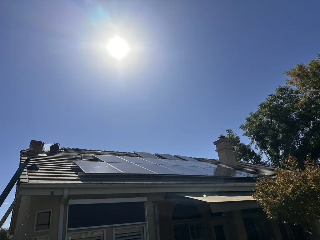Solar panels installed on a house roof under a bright sun with a clear blue sky and a tree on the right side.