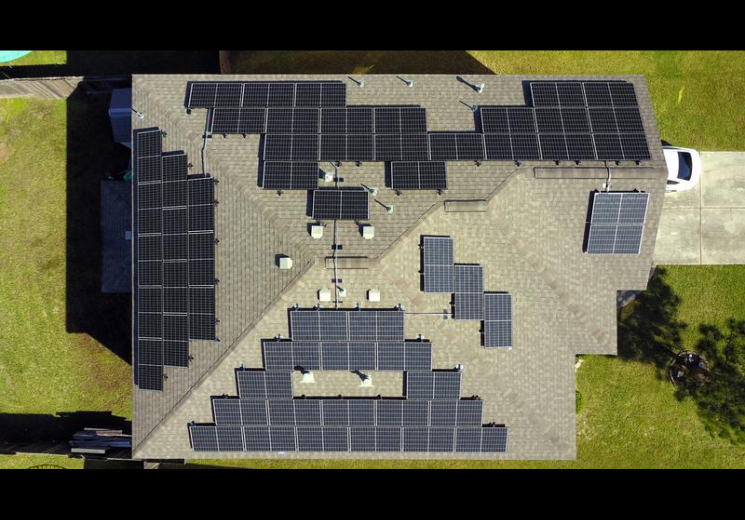 Aerial view of a house with multiple solar panels installed on the roof, driveway, and surrounding green lawn.