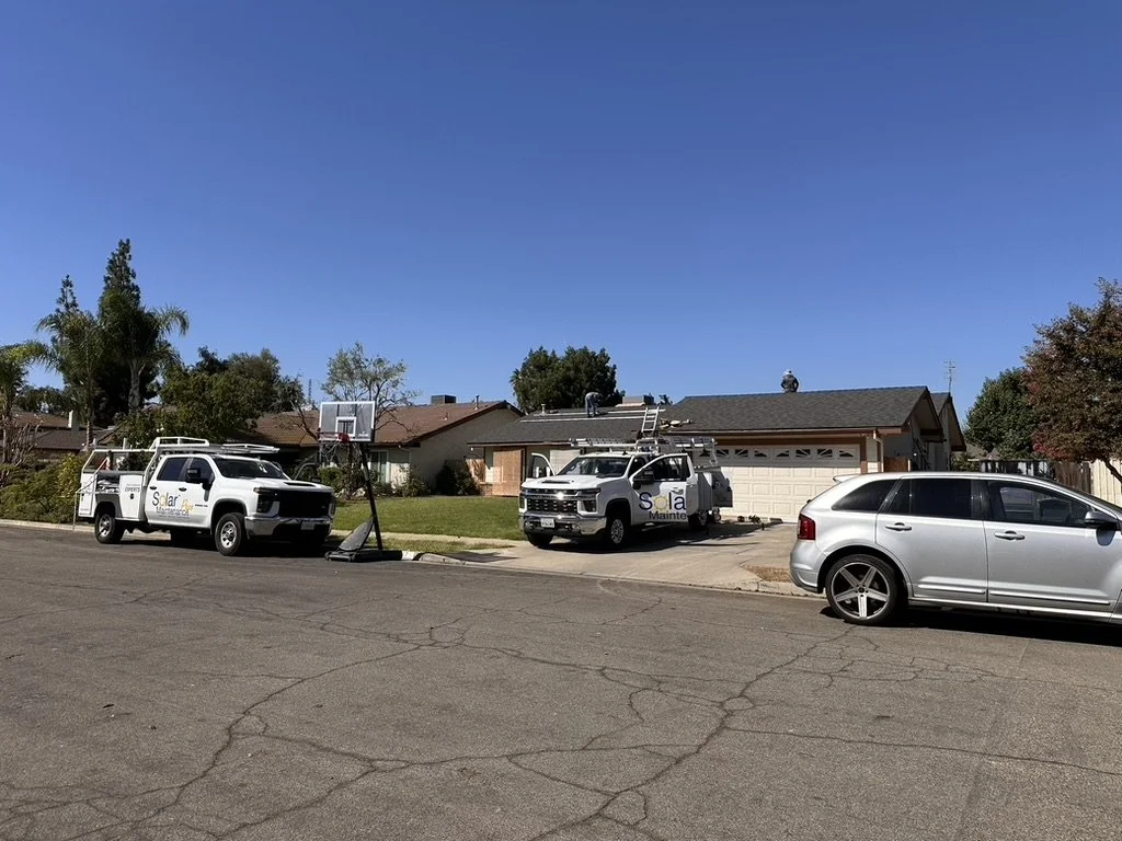 Two white service trucks with ladders parked in front of a suburban house, with a silver car parked on the street. The house has a garage, a basketball hoop, and is surrounded by trees and other houses. The sky is clear and blue.