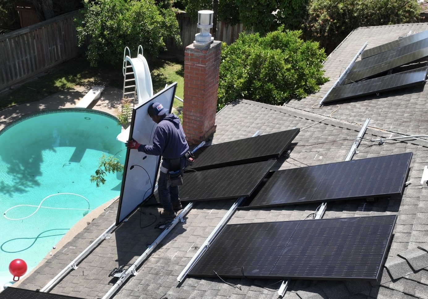 A person installing or maintaining solar panels on a residential roof near a swimming pool, with trees and landscaping in the background.