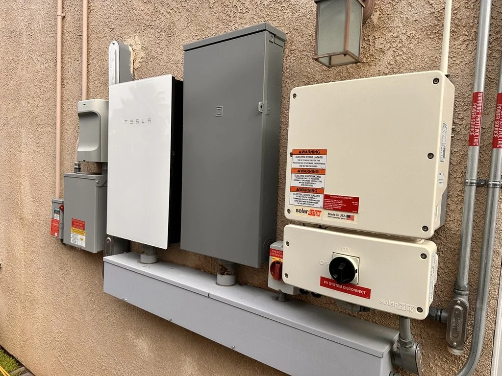 Photograph of electrical components and solar power equipment mounted on an exterior wall, including a Tesla inverter, electrical panels, and warning labels.