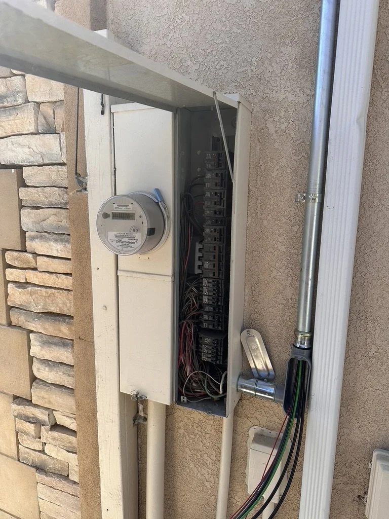 Partial view of an outdoor electrical meter box with wiring and a digital meter attached, mounted on a stucco wall next to a brick wall and a metal outdoor faucet.