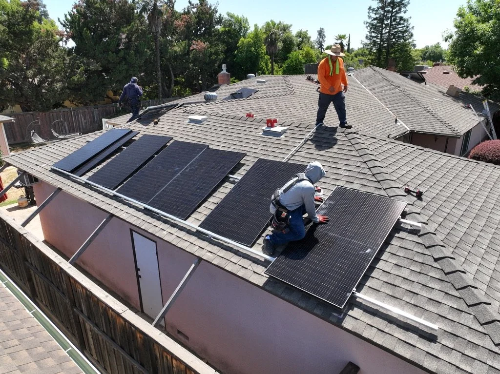 Three workers installing solar panels on the roof of a house, with trees in the background.