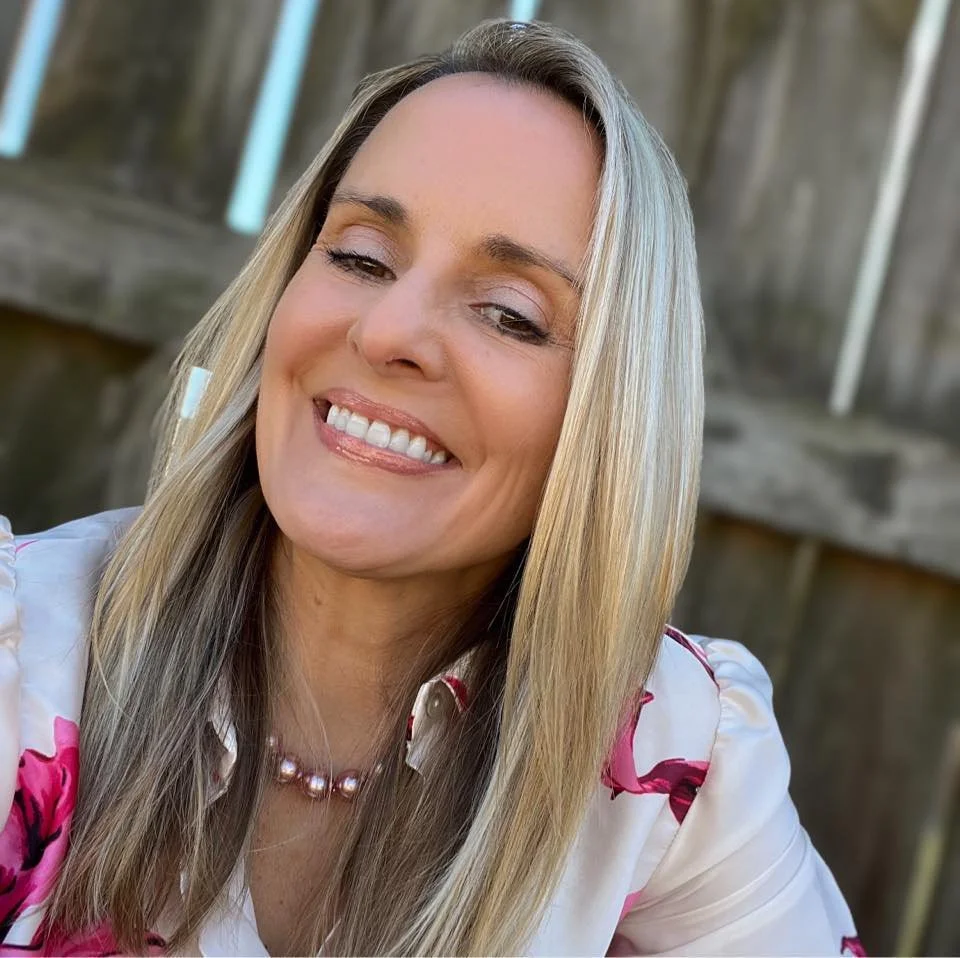 A woman with blonde hair smiling outdoors in front of a wooden fence, wearing a white shirt with pink floral patterns and a pearl necklace.