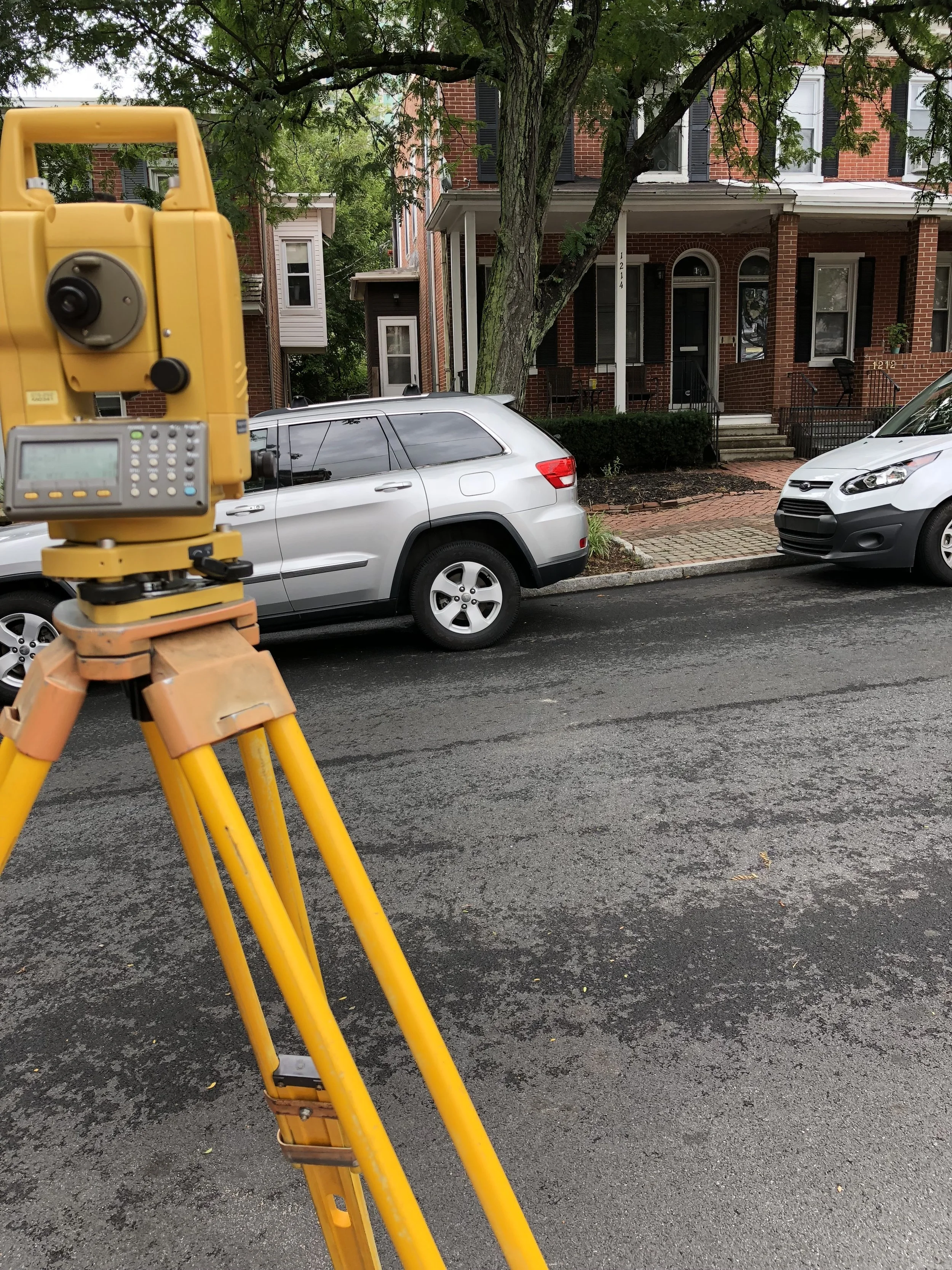A yellow surveying instrument set up on a tripod on a paved street, with a parked silver SUV and a white van in front of a brick residential building with trees. Surveyor conducting mortgage survey in Delaware.
