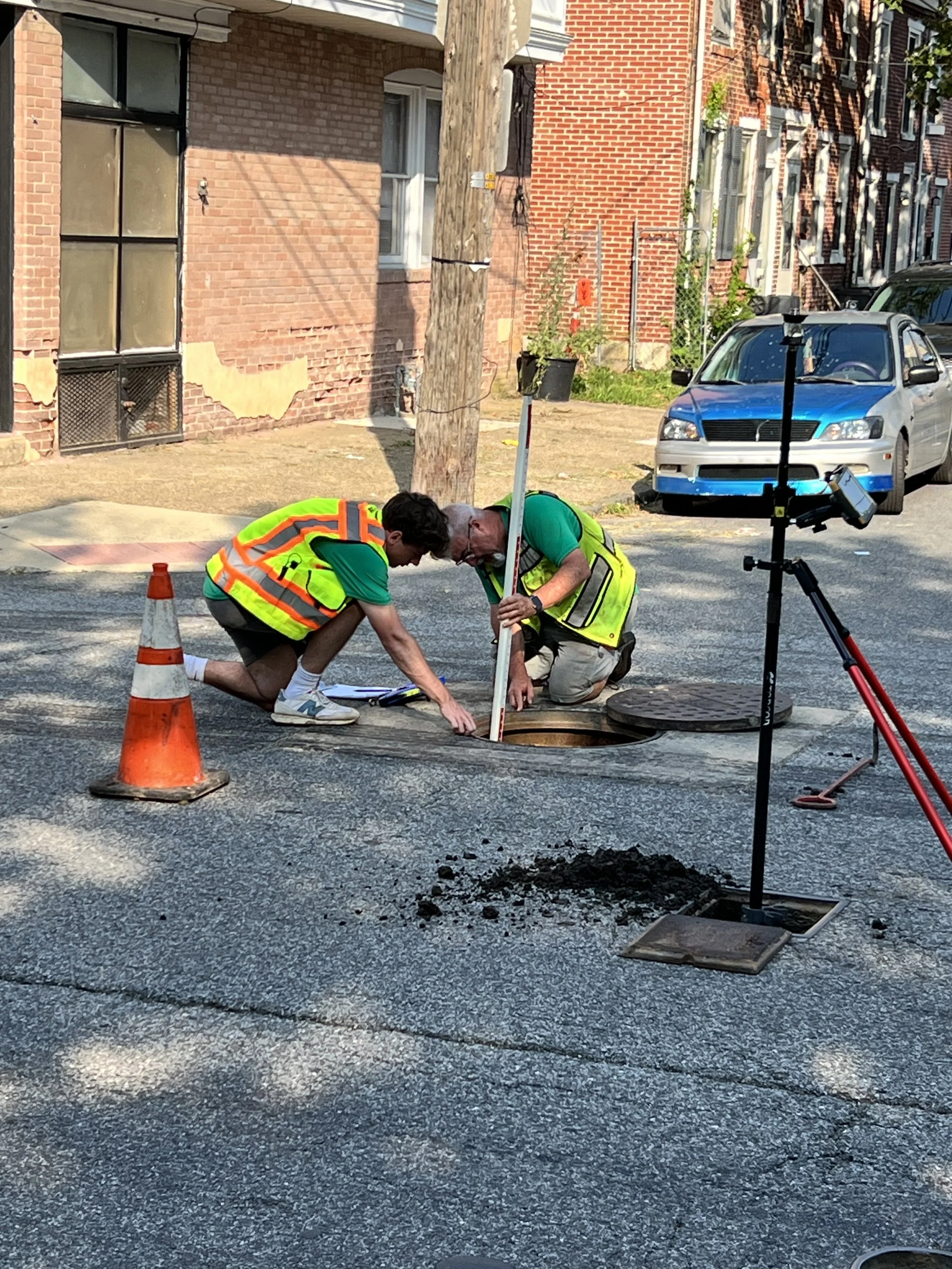 Two workers in yellow safety vests are inspecting an open manhole on a city street. One is crouching and the other is kneeling, both looking into the hole. There is a safety cone and a tripod with a camera or sensor nearby, along with a pile of soil