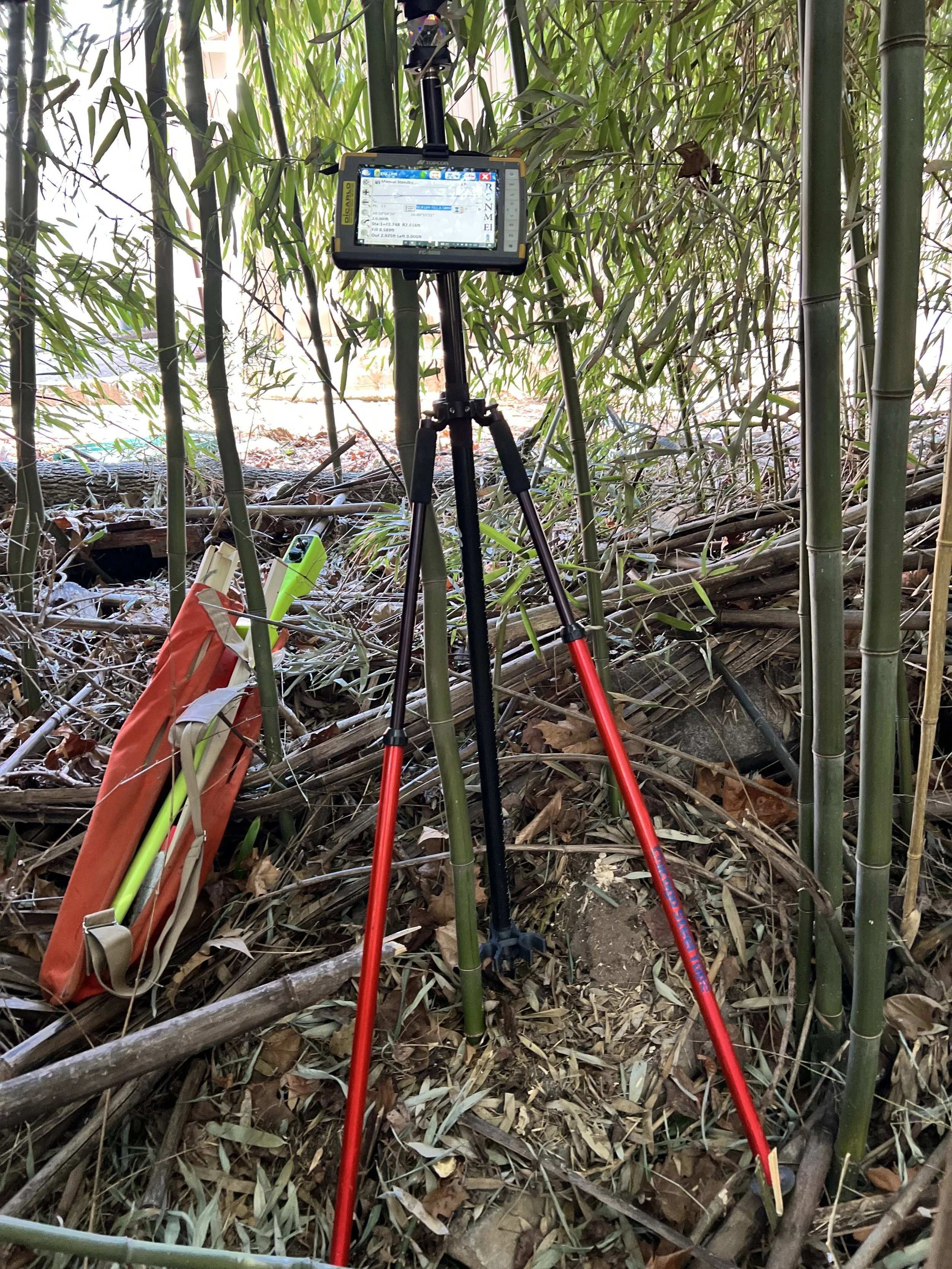 A surveying instrument mounted on a tripod with a digital display screen, set up in a bamboo forest with fallen leaves and branches on the ground.
