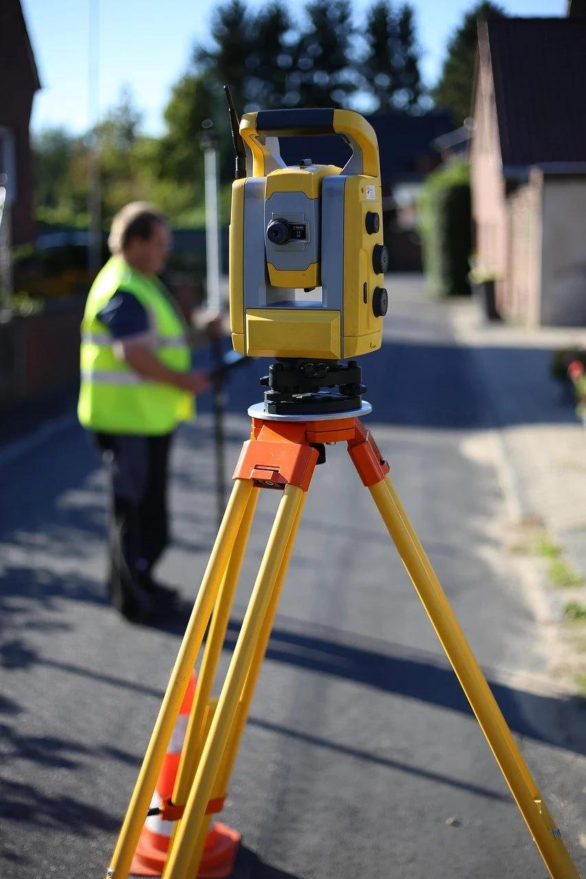 A yellow and gray surveying instrument mounted on an orange tripod on a paved street. In the background, a person wearing a yellow safety vest appears to be working, with houses and trees visible.