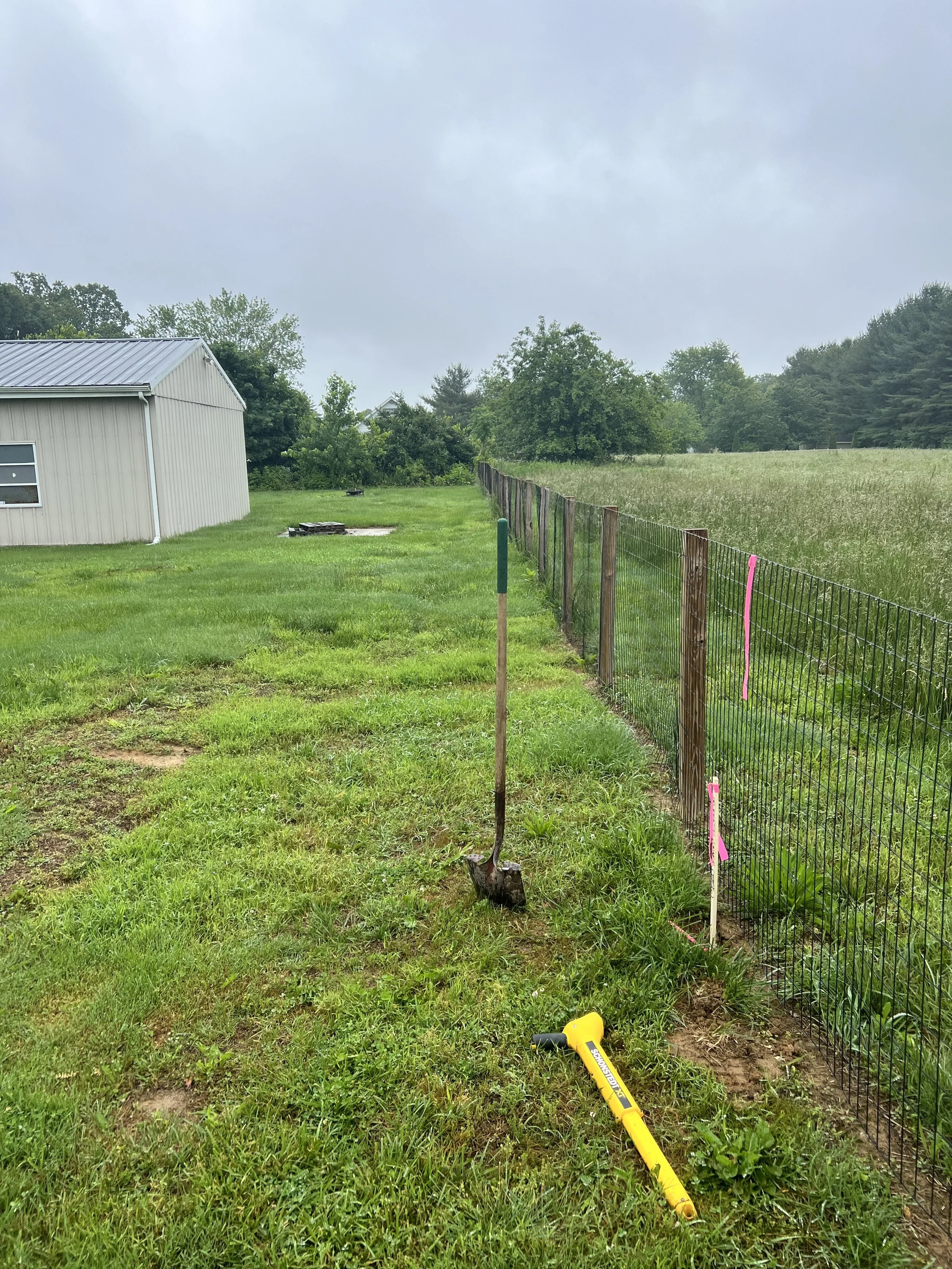A metal shovel and a yellow tool lie on the grass near a new wire fence on a cloudy day. The fence runs along a grassy field, with a shed in the background and trees on the horizon. Property line boundary markers in Delaware.