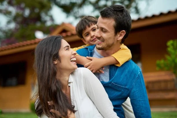 A family of three smiling and playing outside, with a house and greenery in the background.