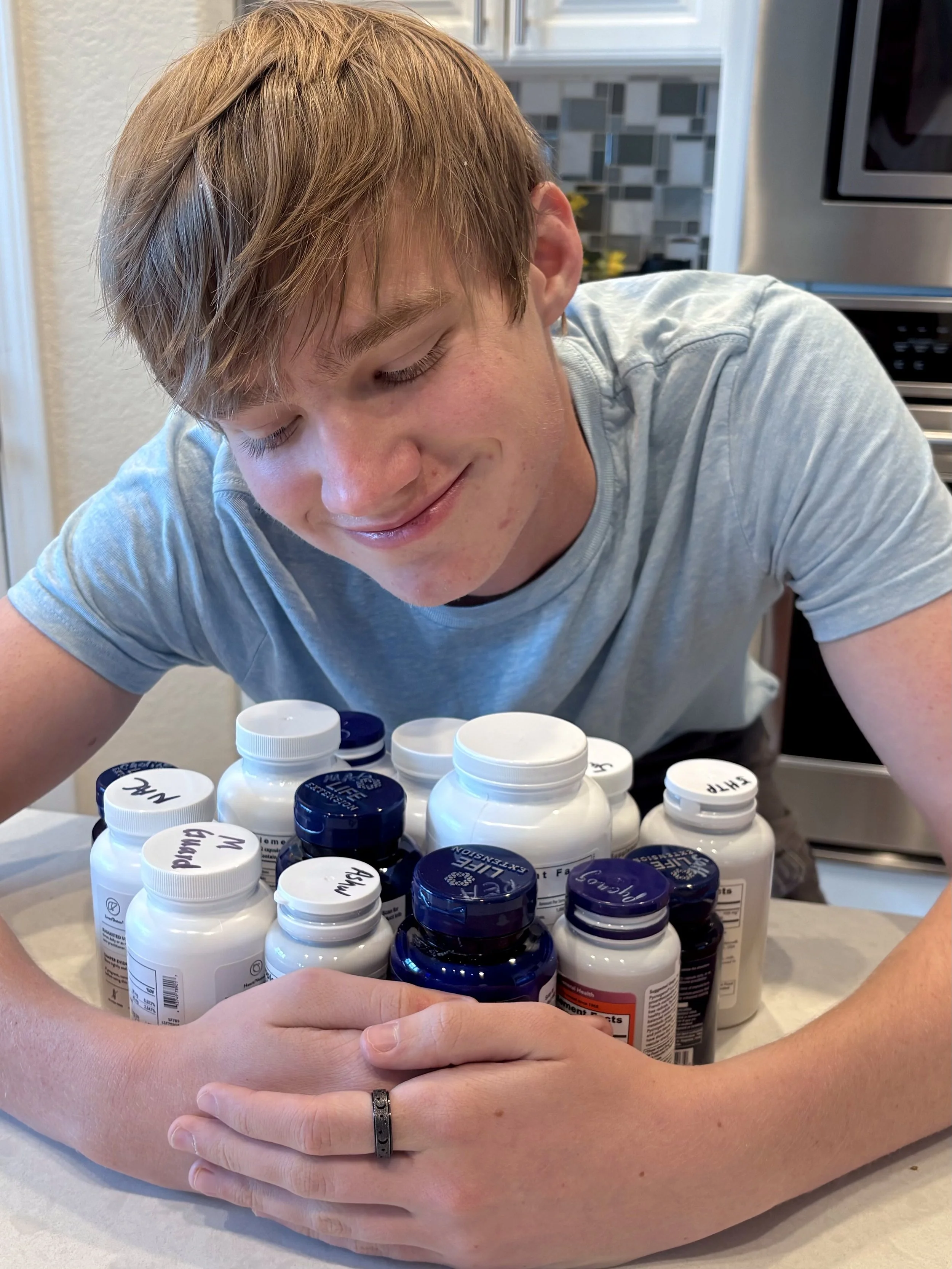 A young man with light skin and short, light brown hair, smiling with closed eyes, is leaning over a collection of white and blue pill bottles on a table, with a kitchen background visible.
