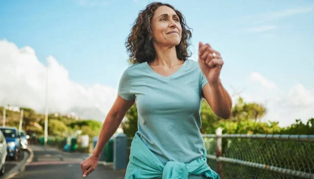 Woman jogging outdoors on a sunny day, wearing a light blue t-shirt and has a sweater tied around her waist.