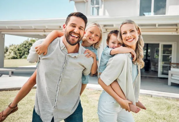 Family of four smiling outside a house, with the father carrying the older daughter on his back and the mother holding the younger daughter, all enjoying a sunny day.