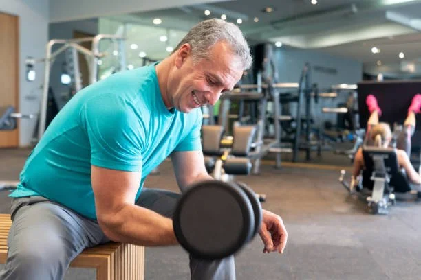 A man in a teal shirt sitting on a bench at a gym, holding a dumbbell, smiling and resting.