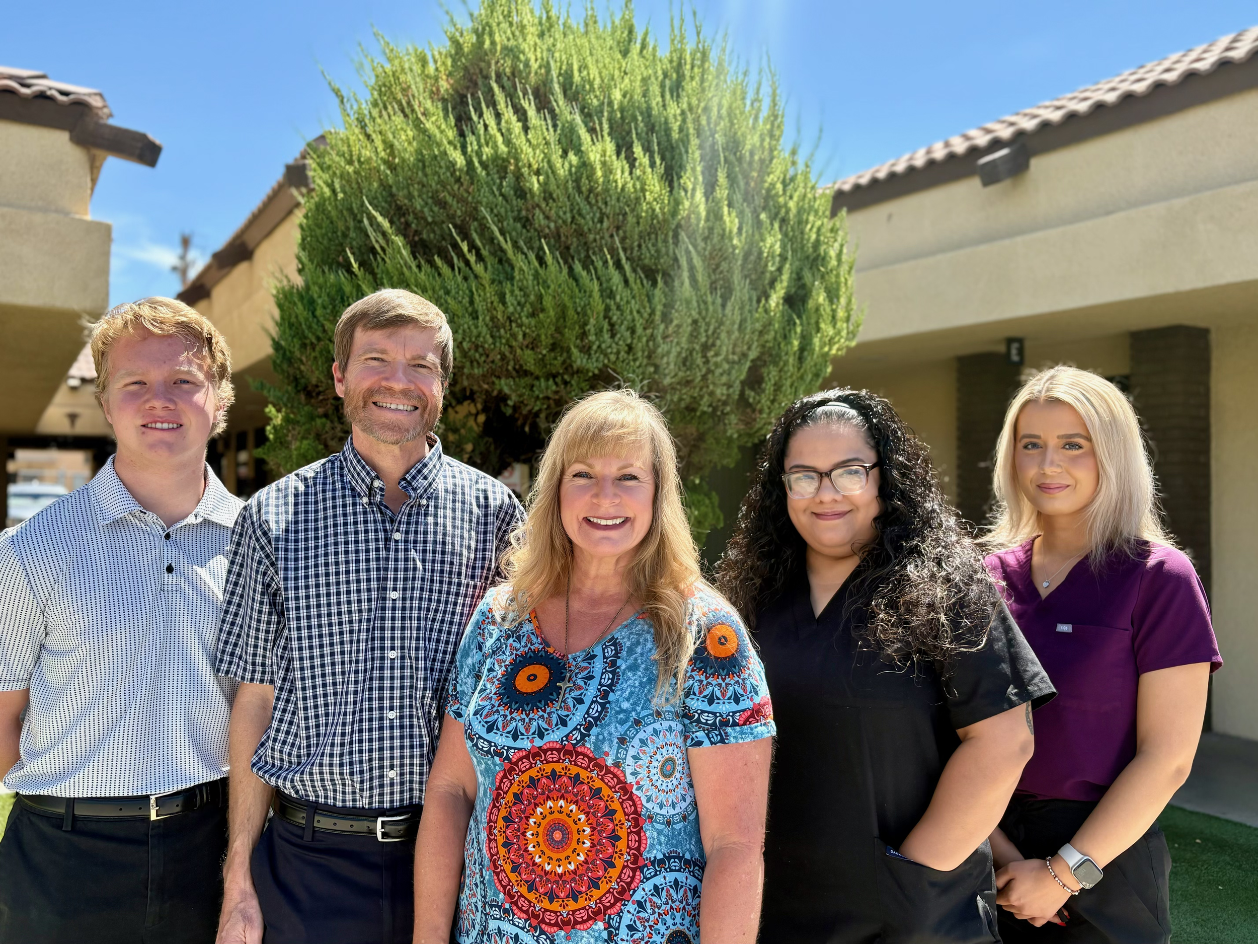 A group of five smiling people standing outdoors in front of a large bush and building, under a blue sky.