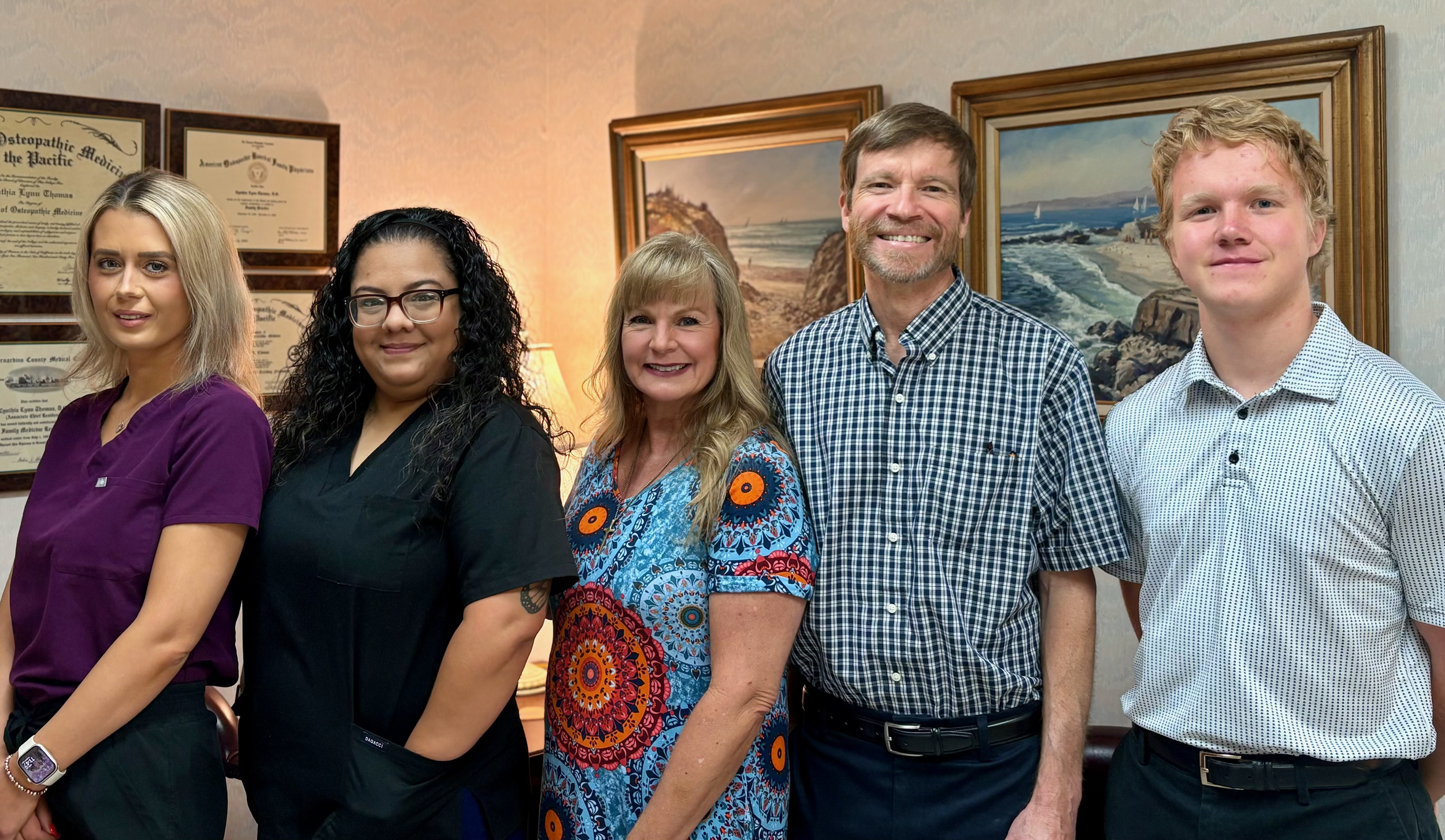 Group of five smiling people standing in a room with framed certificates and ocean-themed paintings on the wall.