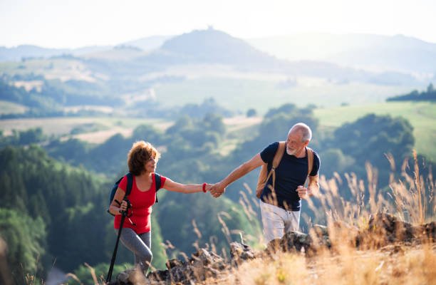 An elderly couple hiking outdoors on a sunny day in a green, hilly landscape, with the woman handing the man a water bottle.