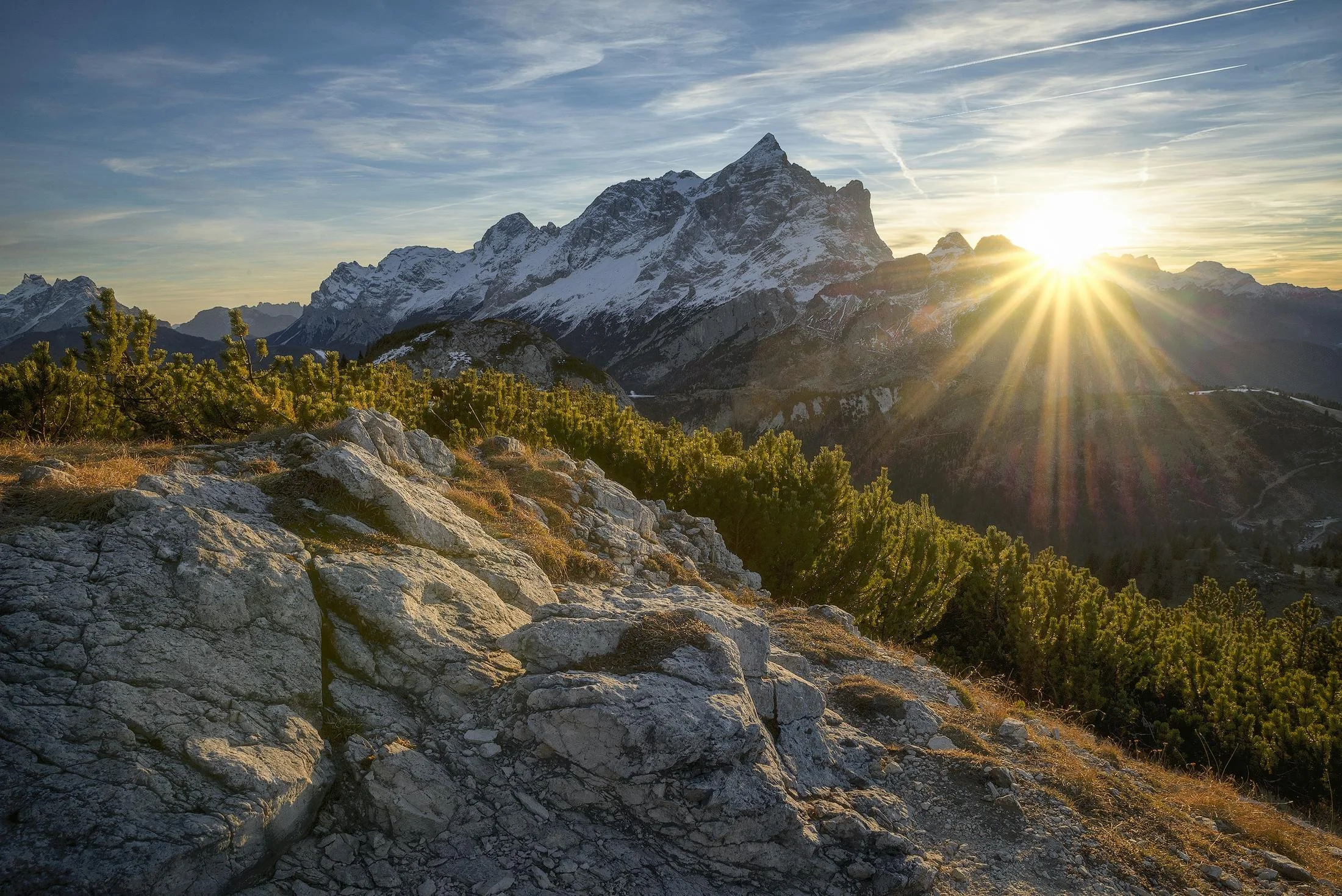 Sunset over snow-capped mountains with rocky foreground and green bushes.