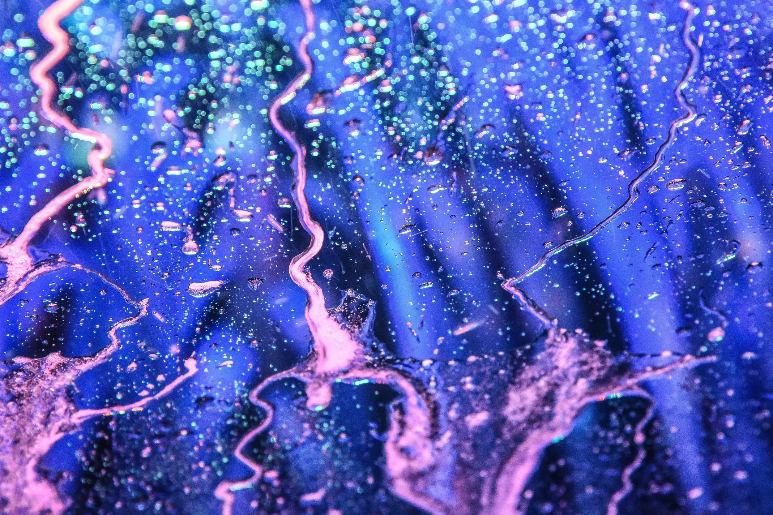 Close-up of shimmering, colorful surface with pink and purple cracks and water droplets, illuminated by vibrant blue, purple, and pink lighting.