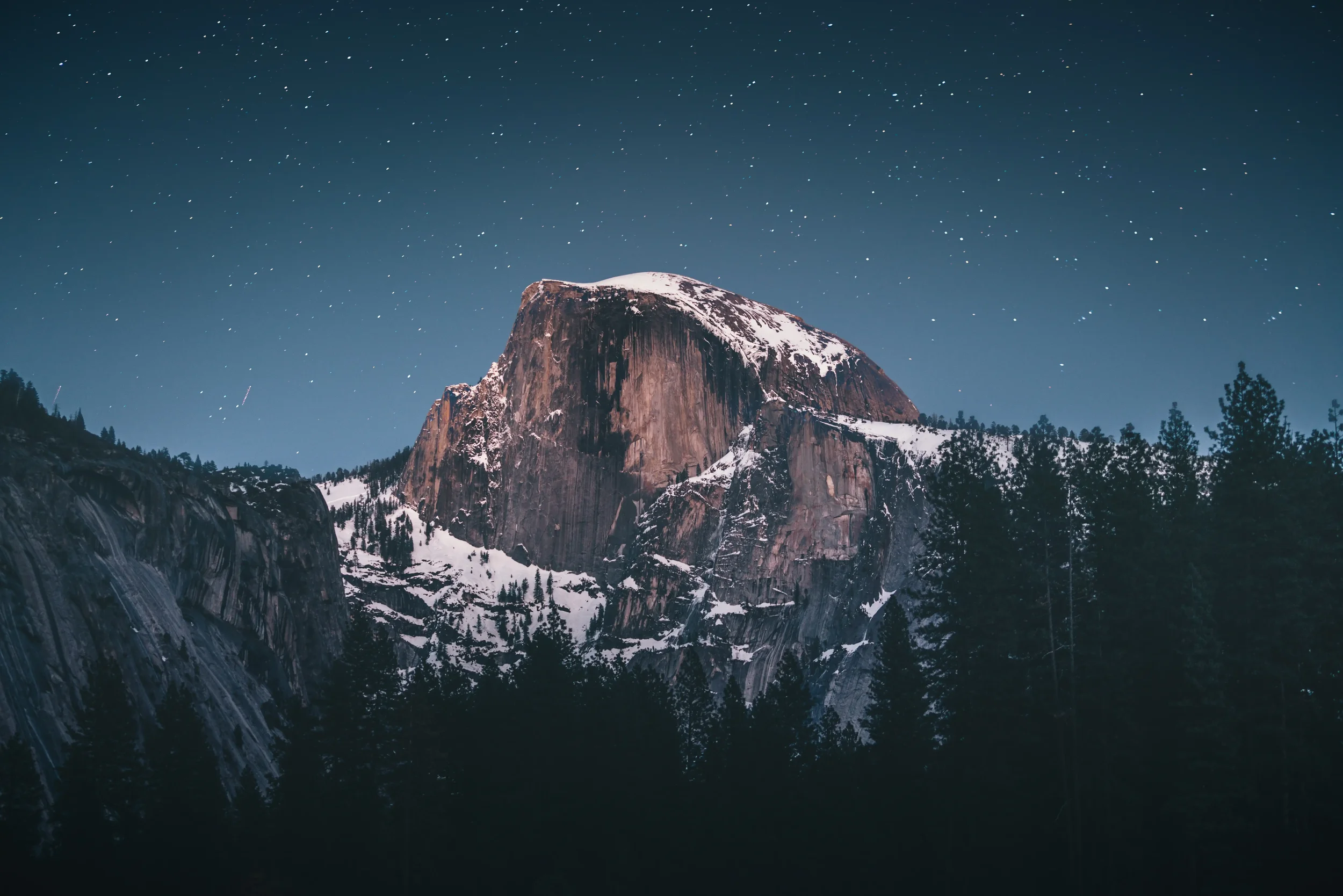 Nighttime view of a mountain with snow on top, dark forest in foreground, and starry sky overhead.
