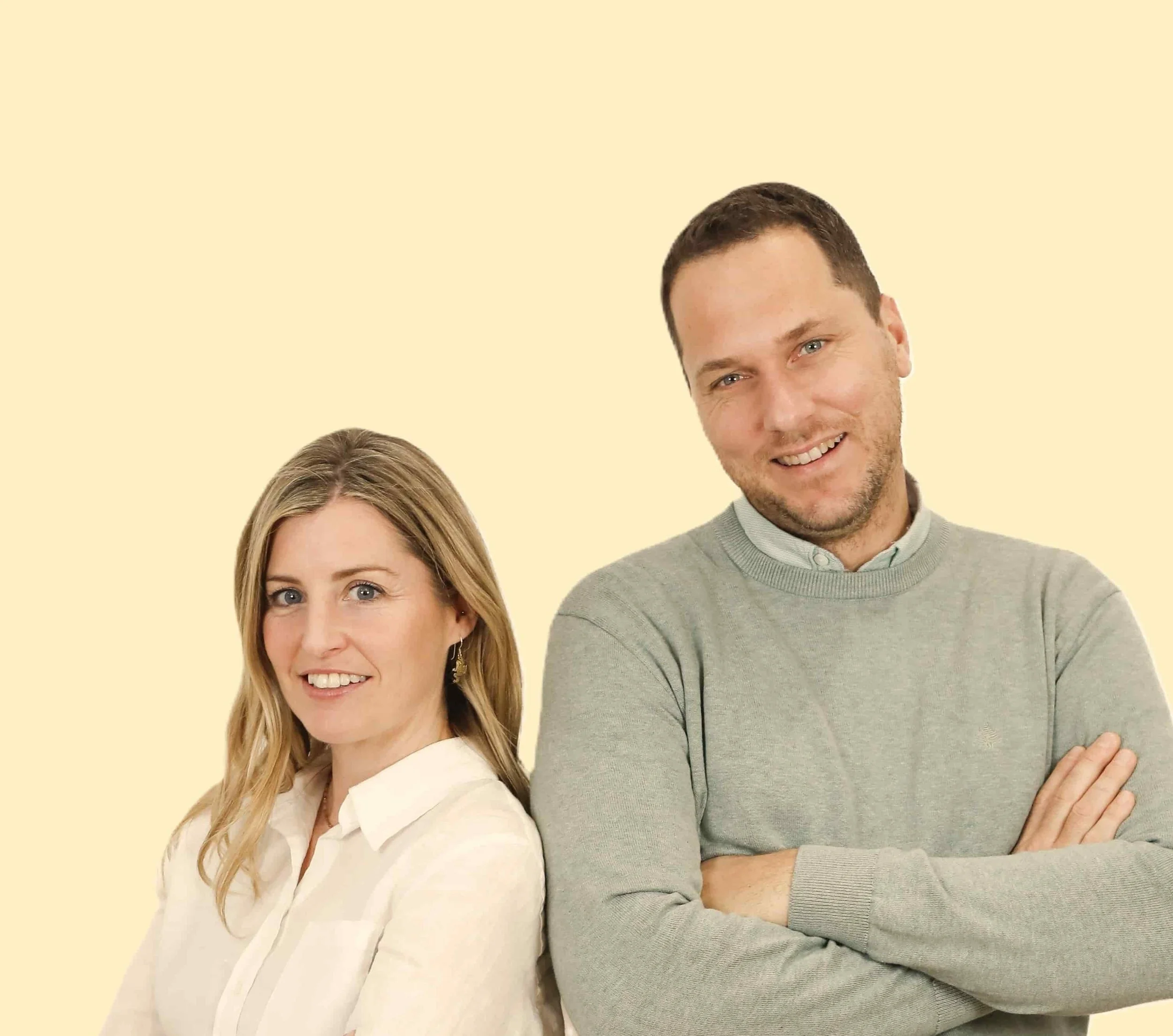 A woman and a man stand back-to-back with arms crossed, smiling against a plain beige background.